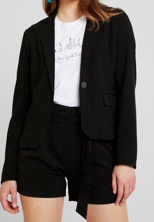 Frau trägt schwarzen Blazer und Shorts mit weißem T-Shirt, das eine Darstellung der Skyline von New York City zeigt.