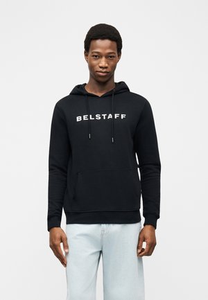 Ung mand iført en sort Belstaff-hoodie med hvidt logotekst og lyseblå jeans, stående mod en ensfarvet lys baggrund.