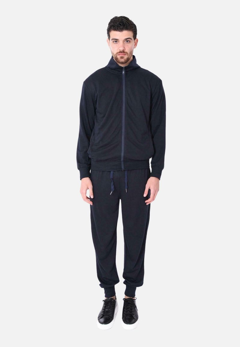 Tuta navy realizzato in tessuto morbido, composto da una giacca con zip e pantaloni comodi con polsini elastici e cordini, abbinato a sneakers nere.