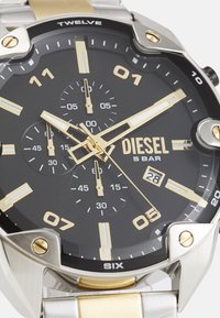 Montre avec cadran noir, accents dorés et argentés, boîtier en acier inoxydable, grands index horaires, sous-cadrans chronographe et affichage de la date.