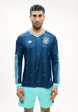 Άνδρας που φοράει μπλε σκούρη φανέλα ποδοσφαίρου με μακριά μανίκια, ανοιχτό μπλε ρίγες και έμβλημα, συνδυασμένη με ανοιχτό μπλε σορτς.