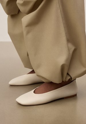 Flade sko med spidse tæer i kremfarve, båret sammen med løsthængende beige bukser, der er samlet ved anklerne, stående på et glat gulv.