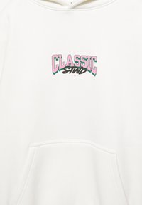 Witte hoodie met een voorvak, met de tekst "CLASSIC STND" in roze en groene retro-stijl letters. Zachte textuur.