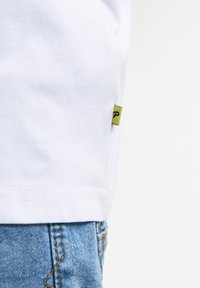 Witte katoenen t-shirt met een zichtbare groene label met een zwarte "P" aan de linkerkant, gecombineerd met blauwe denim shorts.