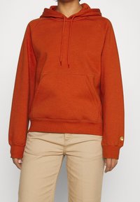 Sudadera naranja con un bolsillo frontal, capucha con cordón, puños y dobladillo acanalados, y detalle del logo en la manga. La textura de la tela parece suave.