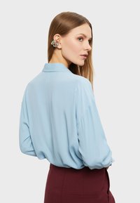 Světle modrá halenka s dlouhým rukávem a límečkem, s uvolněným střihem a knoflíkovými manžetami, spárovaná s vínovými kalhotami. Hladká textilie.