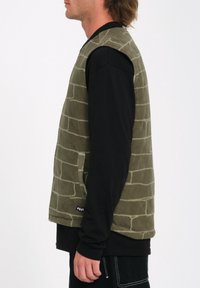 Chaleco texturizado color verde oliva con un patrón de ladrillos, superpuesto sobre una camisa negra de manga larga. Cuenta con bolsillos laterales y una etiqueta de marca.