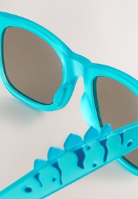 Aqua-blaue Sonnenbrille mit verspiegelten Gläsern; das Gestell zeichnet sich durch ein verspieltes Design mit spitzen Akzenten entlang der Bügel und einer glatten Oberfläche aus.