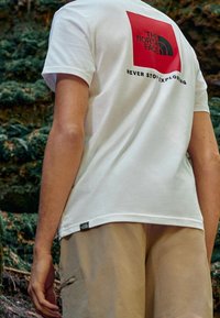 Bílý bavlněný tričko s červeným čtvercovým logem The North Face a textem "NEVER STOP EXPLORING" na zadní straně, v kombinaci s béžovými šortkami.