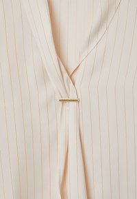 Blusa color pesca chiara con righe verticali dorate, caratterizzata da un design avvolgente e un accento a barra dorata. Texture liscia e lucentezza sottile.