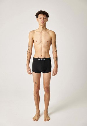 Junger Mann mit Tattoos steht barfuß in schwarzen Snocks-Boxershorts vor einem einfarbigen hellen Hintergrund.