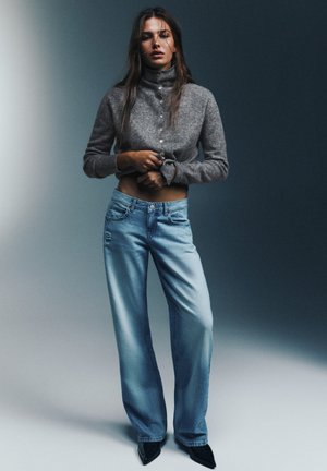 Femme portant un pull court gris à boutons et un jean large bleu clair, debout devant un fond dégradé bleu-gris.