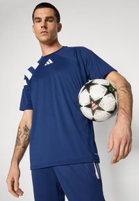 Camisa deportiva azul marino con franjas laterales blancas, combinada con pantalones a juego. El modelo sostiene un balón de fútbol en blanco y negro con detalles gráficos.