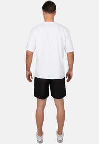 Maglietta bianca in cotone con maniche corte, abbinata a pantaloni corti neri e sneakers bianche, vista dal retro. Design semplice e funzionale.