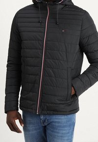 Svart vadderad jacka med dragkedja framtill, har en huva, kontrasterande röda och blå ränder samt sidofickor. Texturerad quiltad design.