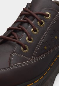 Zapato de cuero marrón oscuro con cordones marrones, suela con costuras amarillas, ojales metálicos, costuras blancas y diseños de flores en relieve en el lateral.