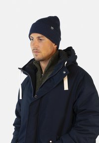 Marineblauwe geïsoleerde jas met een capuchon, witte trekkoorden en daaronder een groene gebreide trui. Een bijpassende marineblauwe gebreide beanie maakt de look compleet.