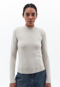 Pitkähiha beige paita korkealla kaula-aukolla, jossa on pystysuoraa ompelua ja slim fit -malleja, yhdistettynä farkku alaosaan.