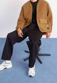 Giacca bomber in pile marrone con chiusura a zip, due tasche frontali, pantaloni neri e sneakers bianche. Seduto su una sedia girevole nera.