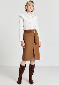 Gonna marrone con uno spacco frontale e decorazioni circolari sulla cintura. Blusa bianca con dettagli arricciati. Stivali in camoscio marroni al ginocchio.