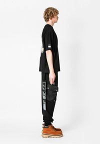 Camiseta negra de gran tamaño con un gráfico en la parte posterior, combinada con pantalones cargo negros que tienen bolsillos y estampados de texto, y botas marrones con cordones.