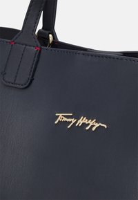 Gros plan sur un sac à main en cuir marine foncé montrant le logo doré de Tommy Hilfiger et l'attache du poignet renforcée par des coutures.