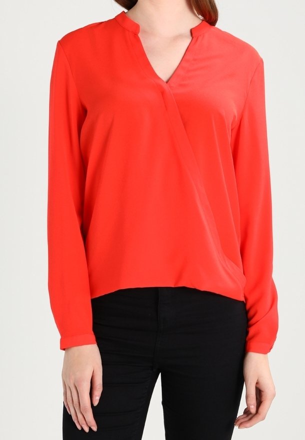 Femme portant un chemisier rouge vif à manches longues avec un col en V et un pantalon noir, debout devant un fond clair.