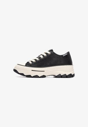 Zapatilla de cuero negra con cordones blancos, suela gruesa beige, acentos de goma negra y una superficie texturizada. Presenta un logo en el talón.