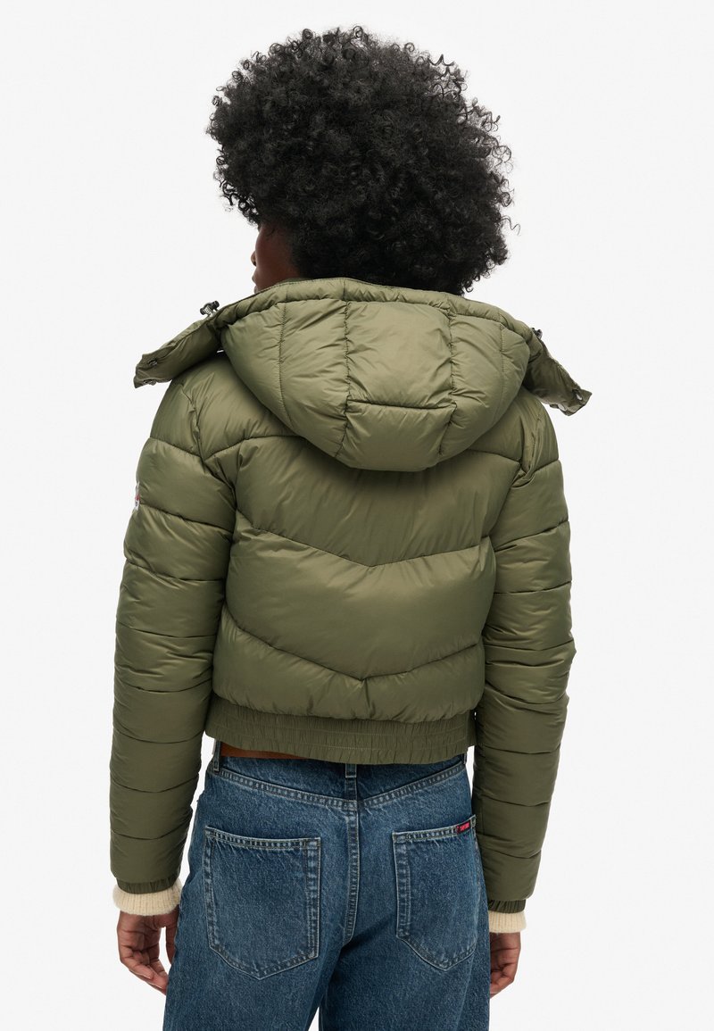 Olivengroene pufferjack met een capuchon, met een doorgestikt ontwerp, elastische manchetten en een cropped lengte. Draagt men met high-waisted jeans.