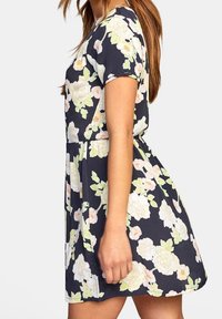 Robe courte florale avec un fond bleu marine. Elle présente des manches courtes, un corsage ajusté et une jupe évasée. Fleurs aux teintes crème et vert.