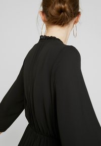 Svart klänning med långa ärmar, med smockad midja och spetsdetaljer vid halsringningen. Slät textur och flödande design, diskreta veck.