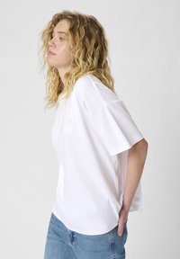 White cotton t-shirt with short sleeves, relaxed fit, featuring a small embroidered logo on the chest. Paired with light blue denim jeans.