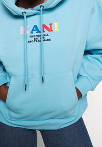 Sudadera azul claro hecha de una tela suave, con un bolsillo frontal tipo canguro y cordones. Gráfico "KANI" en letras coloridas en el pecho.