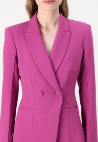 Blazer fuchsia à double boutonnage avec des revers aigus, des coutures latérales et des détails de couture subtils. Fabriqué à partir d'un tissu lisse et extensible.