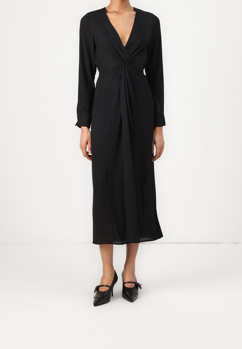 Robe noire à manches longues avec un décolleté en V profond et un détail de taille torsadé, présentant une silhouette fluide et une coupe jusqu'à la cheville. Associée à des talons noirs.