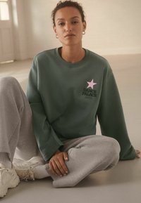 Sudadera verde con una estrella rosa y el texto negro "DANCE PALACE" en el pecho, combinada con pantalones de chándal grises y zapatillas blancas.
