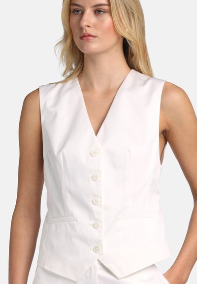 Blondína v bielom bezrukávovom vestu s výstrihom do V, zapínanom na gombíky, s dvoma prednými vreckami, na jednoduchom svetlom pozadí.