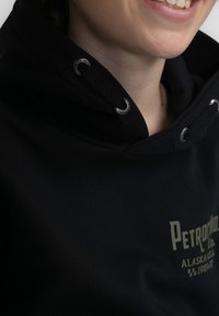 Felpa con cappuccio nera con occhielli argentati, caratterizzata da un logo stampato in verde chiaro e un tessuto testurizzato. Dettaglio di cucitura visibile vicino al colletto.