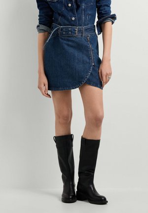 Modelo con falda vaquera azul oscuro con cinturón, chaqueta vaquera a juego y botas de cuero negras hasta la rodilla sobre un fondo liso.