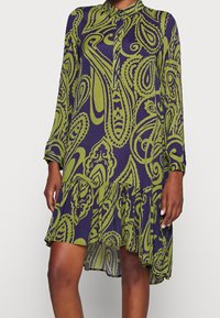 Långärmad klänning i mörklila med grönt paisleymönster, med krage som knäpps framtill och en flerlagers, volangkant. Slät tygkvalitet.