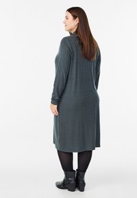 Tumši zaļa adīta kleita ar cīpu rakstu, garām piedurknēm un taisnu griezumu, apvienota ar melniem potīšu zābakiem.