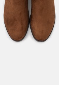Bottines marron en daim avec bouts arrondis, présentant une texture lisse et une semelle épaisse en caoutchouc noir. Design simple sans ornements.