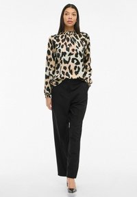 Woman wearing a beige and black leopard print blouse with long sleeves, black tailored pants, and black pointed heels, standing with hands in pockets.