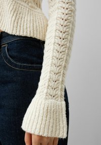 Pull en tricot crème avec un motif texturé de côtes et de cables ; manches évasées et corps ajusté. Porté avec un jean en denim noir.