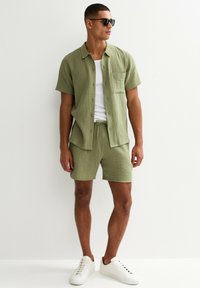 Camicia corta a maniche corte verde chiaro, con texture, e pantaloni abbinati, indossati con una canotta bianca. Completato con sneakers bianche e occhiali da sole neri.
