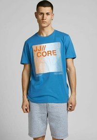 T-shirt in cotone blu con una grafica quadrata testurizzata con "JJ//CORE" in arancione. Maniche corte, collo tondo, abbinato a pantaloni corti grigi.
