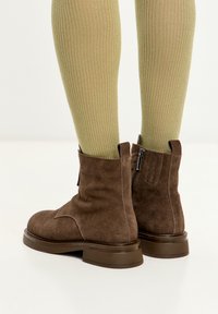 Stivaletti in suede marrone con punta rotonda e tacco basso. Presentano una cerniera laterale e dettagli a coste nella parte superiore, abbinati a calzini a coste verde chiaro.