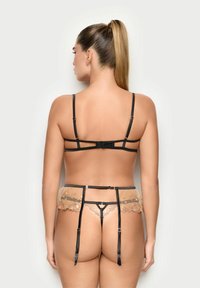 Mujer con coleta larga que lleva lencería negra con tirantes desde atrás, incluyendo un cinturilla de ligas de color beige semitransparente y un tanga a juego.