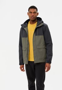 Herrenjacke in dunkelgrün und marineblau, mit Kapuze, Fronttaschen und Druckknöpfen. Weicher Stoff, mit Farbkontrasten auf Brusthöhe.