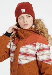Een bruine corduroy jas met een patroon op de borst en schouders in roze en room. Een roestkleurige beanie met een logo-label ligt op krullend haar.